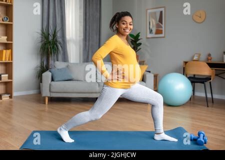 Fröhliche junge, schwanger, schwarze Sportlerin mit großem Bauch, die Beinübungen macht und Yoga praktiziert Stockfoto