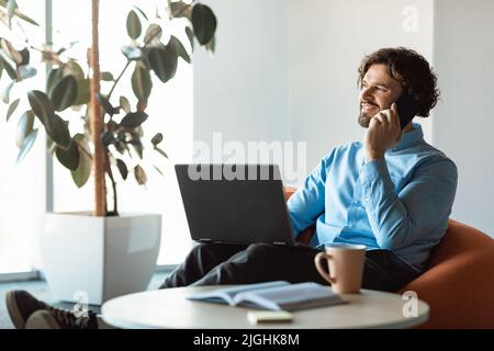 Lächelnder, reifer Geschäftsmann, der mit dem Handy spricht, einen Laptop benutzt, in einem Sitzsack im Firmenbüro sitzt, Platz zum Kopieren Stockfoto