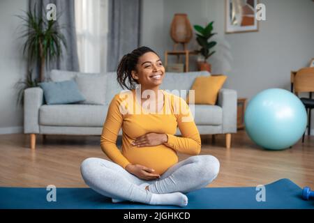 Zufriedene Millennial schwanger afroamerikanische Dame berührt großen Bauch fühlt Bewegung des Babys, Yoga zu üben Stockfoto