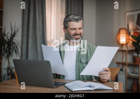 Lächelnder kaukasischer älterer Mann mit Bart arbeitet auf einem Laptop mit Dokumenten im Wohnzimmer Stockfoto