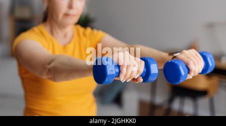 Sportlich gealterte Frau hebt Hanteln auf, trainiert im Wohnzimmer, arbeitet an der Armstärke, selektiver Fokus, Panorama Stockfoto