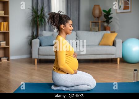 Zufriedene ruhige junge, schwanger afroamerikanische Frau sitzt auf der Matte, praktiziert Yoga und Meditation Stockfoto