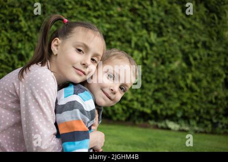 Porträt von charmanten lächelnden jüngeren Bruder und ältere Schwester, sitzen zusammen im Park, umarmt. Happy Lifestyle Kids. Freundschaftliche Beziehungen zwischen Stockfoto