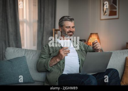 Fröhlicher kaukasischer reifer Mann mit Bart sieht auf Laptop, hält Kreditkarte und Telefon Stockfoto