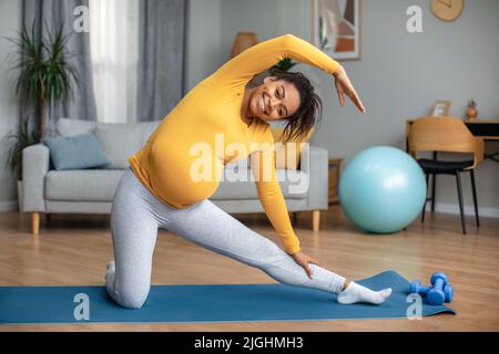 Glücklich lächelnd junge, schwanger schwarze Frau, die Yoga praktiziert und den Körper im Wohnzimmer auf einer Matte dehnt Stockfoto