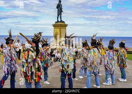 Die Shropshire Bedlams und Martha Rhodens Tänzer Tuppenny Dish Morris treten mit einer Bronzestatue von Captain Cook im Hintergrund in Whitby, Großbritannien, auf. Stockfoto