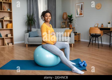 Glückliche junge schwanger afroamerikanische Frau berühren großen Bauch, genießen Sie die Übung auf Fitness-Ball Stockfoto