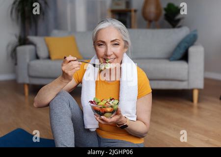 Sport- und Ernährungskonzept. Glücklicher Senior femael, der sich zu Hause auf einer Yogamatte mit frischem Gemüsesalat lehnte Stockfoto