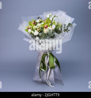 Blumenstrauß in hellgrüner Farbe, der in hellem Blumenpapier gehüllt ist, steht auf blauem Hintergrund. Zartes Bouquet von weißen Rosen, eustoma, gre Stockfoto