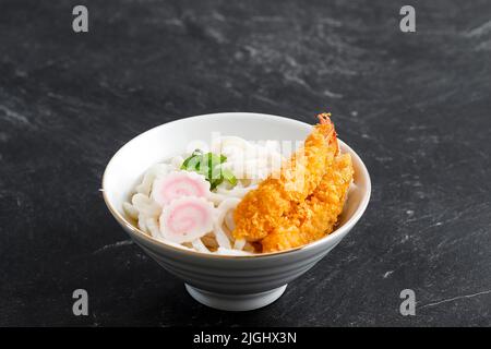 Japanische Tempura Udon-Nudeln mit Garnelen-Tempura und Narutomaki-Topping auf weißer Schale über schwarzem Marmortisch Stockfoto