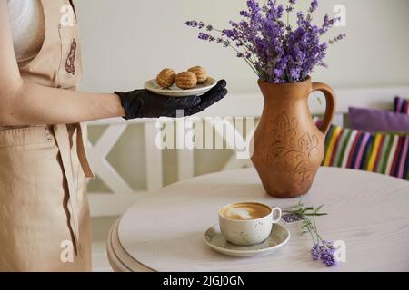 Kellner serviert vegane, natürliche Nüsse, Süßigkeiten ohne Zucker. Tasse Cappuccino auf einem Holztisch im Café. Natürliche Süßigkeiten. Kaffeepause. Lavendeldekor Stockfoto