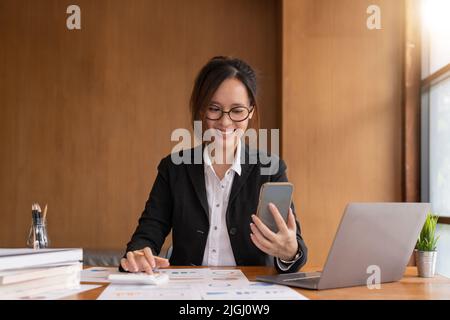 Asiatische Frau hält Smartphone, während mit Taschenrechner für Business-Finanzbuchhaltung berechnen Geld Bank Darlehen Miete Zahlungen verwalten Aufwendungen Stockfoto