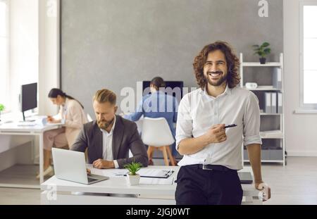 Porträt eines glücklichen jungen Geschäftsmanns, der im Büro steht, den Stift hält und lächelt Stockfoto