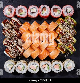 Asiatisches Essen ist fest. Nahaufnahme verschiedener Sushi-Arten, die auf dunklem Hintergrund serviert werden. Menü Sushi mit Nigiri, Maki, Uramaki auf schwarzem Teller. Japanisches Restaurant Stockfoto