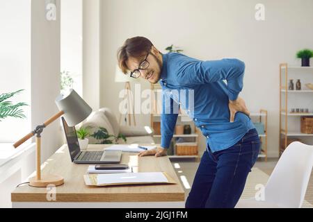 Der männliche Büroangestellte hat starke Rückenschmerzen, die durch längeres Sitzen in falscher Position verursacht werden. Stockfoto