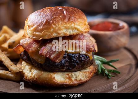 Nahaufnahme von Speck-Cheeseburger auf getoastetem Brötchen mit pommes auf einer Holzplatte Stockfoto