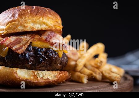 Nahaufnahme von Speck-Cheeseburger auf getoastetem Brötchen mit pommes auf einer Holzplatte. Schwarzer Hintergrund mit Kopierbereich Stockfoto