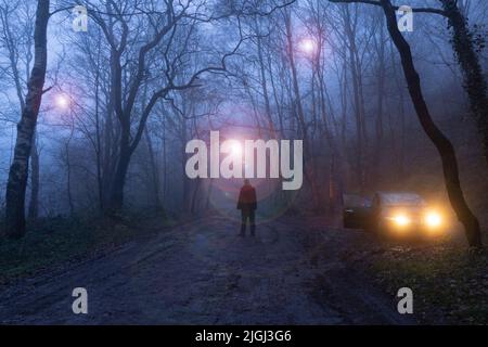 Ein Mann, der neben dem Auto steht und glühende UFOs durch Bäume schweben sieht. In einem gruseligen Wald in einer nebligen Winternacht. Stockfoto