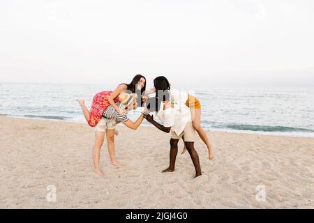 Zwei multirassische Männer kämpfen, während sie ihre Freundinnen am Strand huckepack Stockfoto
