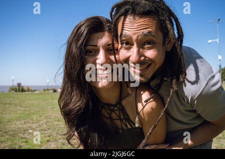 Porträt eines jungen Latiners mit Dreadlocks und einer kaukasischen Frau, die sich umarmen, gemeinsam am Strand im Freien, sich nach vorne lehnen und die Kamera betrachten Stockfoto