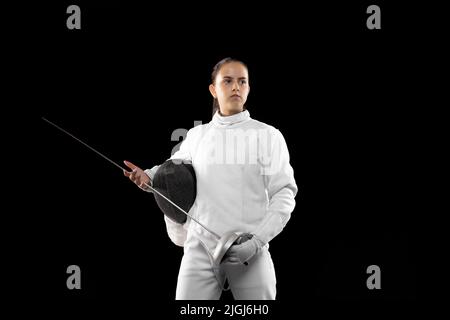 Junge seriöse Mädchen, Anfänger Fechter im weißen Fechtkostüm stehend mit Rapier und Maske isoliert auf dunklem Hintergrund. Sport, Jugend, Erfolge Stockfoto