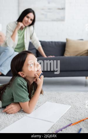 Verträumtes Mädchen liegt auf dem Teppich und denkt an verschwommenes Kindermädchen, das auf dem Sofa sitzt Stockfoto