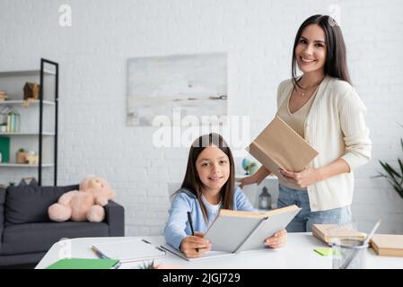Glückliches Mädchen macht Hausaufgaben und hält Buch in der Nähe lächelnde Babysitter Blick auf die Kamera Stockfoto