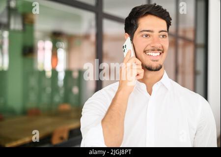 Porträt eines jungen hispanischen Büromitarbeiters im formellen Hemd, der in Innenräumen telefoniert, ein Mitarbeiter mit weißem Kragen ruft sein Smartphone an, diskutiert Pläne, ein Mann schaut mit zufriedenem Gesichtsausdruck beiseite Stockfoto