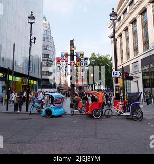 Rikschas aka Pedicabs aka Tuk-Tuks aka Bike Cab am Rande des Leicester Square, vor der Schweizer Kantonsplagge. London. Stockfoto