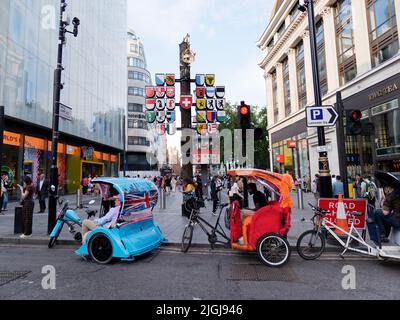 Rikschas aka Pedicabs aka Tuk-Tuks aka Bike Cab am Rande des Leicester Square, vor der Schweizer Kantonsplagge. London. Stockfoto