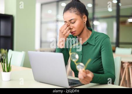 Müde junge afroamerikanische Mitarbeiterin nahm die Brille ab und massiert den Nasenrücken und leidet unter schweren Kopfschmerzen Migräne. Müde Freiberuflerin Frau überwältigt von Computer-Arbeit Stockfoto