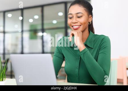 Positive junge afroamerikanische Frau, die im Loft-Stil auf dem Schreibtisch sitzt und einen Laptop benutzt. Fröhliche Angestellte, die auf der Tastatur tippt, schaut zufrieden auf den Bildschirm Stockfoto