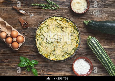 Hausgemachte Zucchini-Pie und Zutaten - Parmesan, Eier und Kräuter auf dem Holzhintergrund, Draufsicht Stockfoto