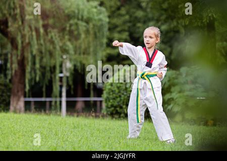 Kaukasisches Mädchen, sieben Jahre alt, im Kimono mit gelbgrünem Gürtel, das Taekwondo im Sommerpark allein während der Coronavirus-Covid-19-Sperre trainiert Stockfoto