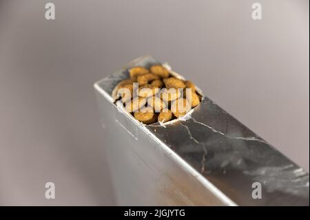 Eine silberne rechteckige Box auf grauem Hintergrund. Tierfutter wird aus dem Loch ausgespült. Trockene, pelletierte Lebensmittel. Tierfutter. Selektiver Fokus. Stockfoto