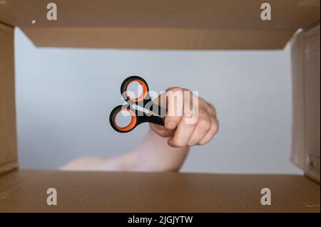 Eine Männerhand legt eine Schreibwarenschere in eine bewegliche Schachtel aus Pappe. Handwerkzeug zum Schneiden von Papier und anderen dünnen Objekten. Ansicht von unten. Innenansicht. Clos Stockfoto