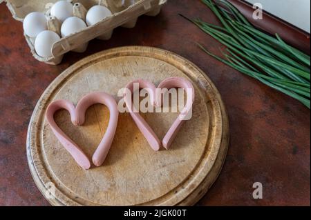 Zwei herzförmige Würstchen liegen auf einem Schneidebrett. Ein Tablett mit Hühnereiern und einem Bund grüner Zwiebeln stehen nebeneinander. Kochen der Mahlzeit. Liebe, Stockfoto