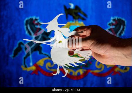 Hexe, die auf einem Besenstiel vor der Flagge des Commonwealth of Pennsylvania fliegt. Mann Hand hält Figur aus Papier geschnitten. Wir feiern die Allerheiligen-Nacht in Stockfoto