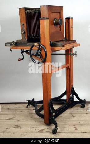 Bälgkamera Kamera (Balgenkamera). Dreiseitiger Sockel aus schwarzem Gusseisen. Ein Rad in jeder Ecke. Lage des lackierten Holzes. Gusseisen-Rad an der Seite, mit Holzgriff, zur Höhenverstellung. Metallschild mit Text 'Century made by Eastman Kodak Co. Successor to Century Camera Co. Rochester NY'. Auf einer kurzen Seite der Platte ist eine „Strebe“ montiert. Textplatte „A. Slewka, Hindenburg O/S Vignelten Halter Dresden 1909 Silber. Medallie“. Die Vorder- und Rückseite der Kamera gleitet auf zwei Zahnstangen, die sich in der Bodenplatte befinden. Im vorderen Teil eine abnehmbare Platte, in der das Objektiv sitzt. Dies Stockfoto