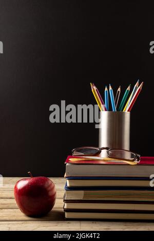 Vertikale Abbildung von Büchern, Gläsern, Buntstiften im Behälter und Apfel auf schwarzer Oberfläche Stockfoto