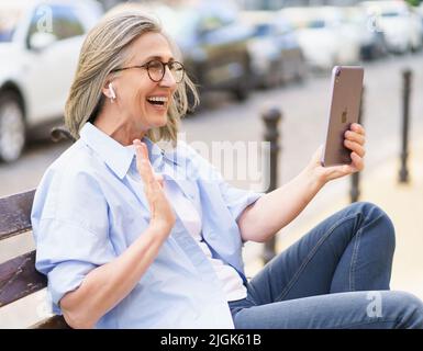 Videoanruf mit digitalem Tablet und Kopfhörern eine reife Frau mit grauen Haaren sitzt auf der Bank in den Straßen der alten europäischen Stadt. Reife Frau in Denim und blauem Hemd im Freien. Stockfoto