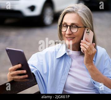 Genießen Sie die Freizeit reife graue Haare Frau sprechen auf dem Telefon Lesen Nachrichten von digitalen Tablet auf der Bank auf den Straßen der alten europäischen Stadt sitzen. Reife Frau nimmt Anruf im Freien. Stockfoto