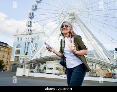 Europäische charmante grauhaarige Frau glücklich genießen Freizeit nach der Arbeit oder auf Reisen, Telefon halten, während Saft auf dem Weg mit Plastikbecher in der Stadt und Riesenrad auf dem Hintergrund. Stockfoto