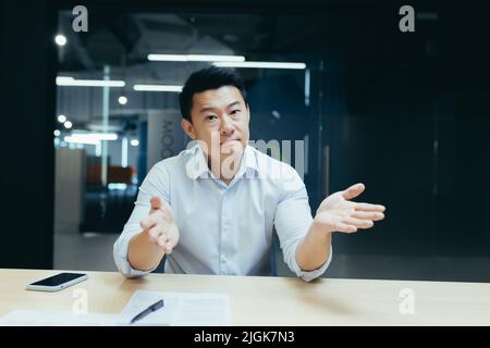 Asiatischer Geschäftsmann bei einem Online-Meeting, hört ernsthaft auf den Gesprächspartner, breitet hilflos die Hände aus, schaut auf die Kamera, Videoanruf Stockfoto