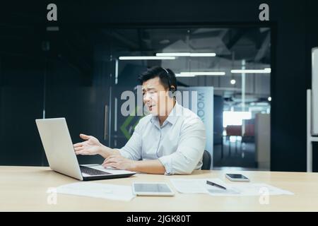 Verärgerter und verärgerter asiatischer Geschäftsmann mit Headset für Videoanruf, Gespräch in Online-Meetings, Blick auf den Laptop-Bildschirm, Mann bei der Arbeit Stockfoto