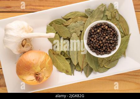 Trocken duftender schwarzer Pfeffer, Lorbeerblatt, Zwiebel und Knoblauch mit weißen Keramikutensilien auf einem Holztisch, Nahaufnahme, Draufsicht. Stockfoto