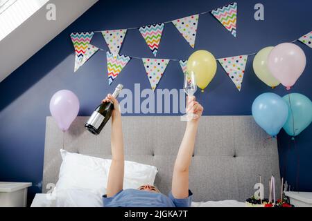 Frau mit erhobenen Armen hält Flasche und ein Glas Champagner auf dem Bett mit festlichem Kuchen im dekorierten Schlafzimmer liegen.Zeit für sich selbst Feier Stockfoto
