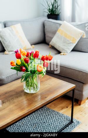 Vase mit frischen Tulpen auf dem Couchtisch mit verschwommenem Hintergrund des modernen gemütlichen hellen Wohnzimmers mit grauem Sofa, grafischen Kissen und grünem pla Stockfoto