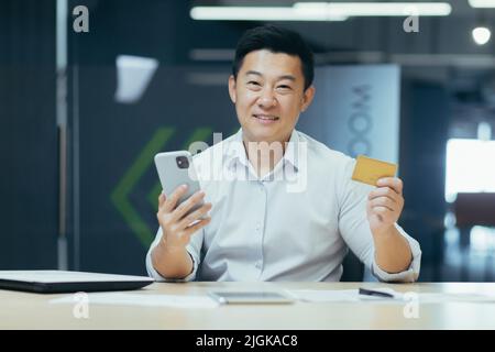 Porträt eines erfolgreichen asiatischen Mannes im Büro, eines Geschäftsmannes, der die Kamera anschaut und lächelt, mit einer Bankkarte und einem Smartphone für das Online-Shopping im Online-Shop Stockfoto