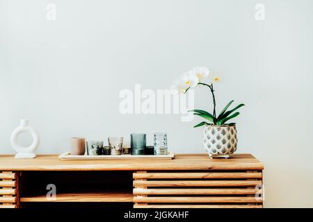 Weiße Orchidee in Blüte und Tablett mit Kerzen auf dem Holzschrank gegen die graue Wand in moderner skandinavischer minimalistischer Wohneinrichtung. Home dekoriert Stockfoto